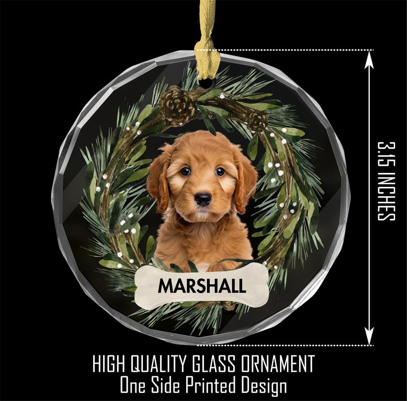 May include: A round, clear glass Christmas ornament with a festive green wreath design. A brown and white puppy is inside the wreath, with the name "MARSHALL" on a bone-shaped tag. The ornament is 3 1/5 inches in diameter.