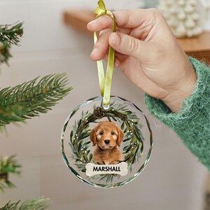 May include: A round glass Christmas ornament with a green wreath and a golden retriever puppy inside. The puppy is looking at the camera and the ornament has the name "MARSHALL" written on it.