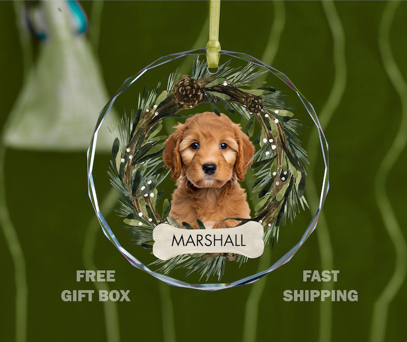May include: A round glass Christmas ornament featuring a brown dog with a green wreath and the name "MARSHALL" on a bone.