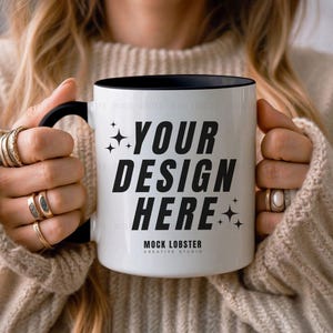 May include: A white ceramic mug with a black rim and handle. The mug has the text "YOUR DESIGN HERE" in black lettering and the words "MOCK LOBSTER CREATIVE STUDIO" in smaller black lettering.