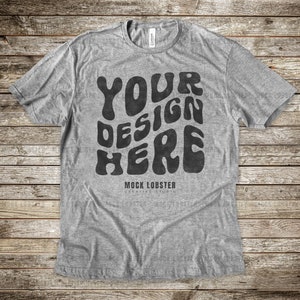 May include: A heather gray t-shirt with a vintage-style graphic that says "YOUR DESIGN HERE" in black. The text is in a wavy, psychedelic font. The shirt is laid flat on a wooden surface.