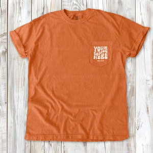 Pode incluir: Uma camiseta laranja queimada com um bolso do lado esquerdo. O bolso tem o texto "YOUR DESIGN HERE" impresso nele.