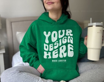 May include: A green hooded sweatshirt with the text "YOUR DESIGN HERE" in white, retro-style lettering. The text is surrounded by a white, wavy design. The sweatshirt is worn by a person sitting on a bed. The text "MOCK LOBSTER CREATIVE STUDIO" is printed below the design.