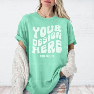 May include: A woman wearing a light green t-shirt with the text "YOUR DESIGN HERE" in white. The shirt is oversized and has rolled up sleeves. The woman is wearing a white cardigan over the shirt and blue jeans.
