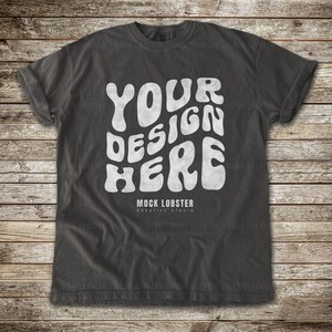 May include: Dark gray t-shirt with the text "YOUR DESIGN HERE" in a white, wavy font. The text "MOCK LOBSTER CREATIVE STUDIO" is below the main text. The shirt is displayed on a wooden background.
