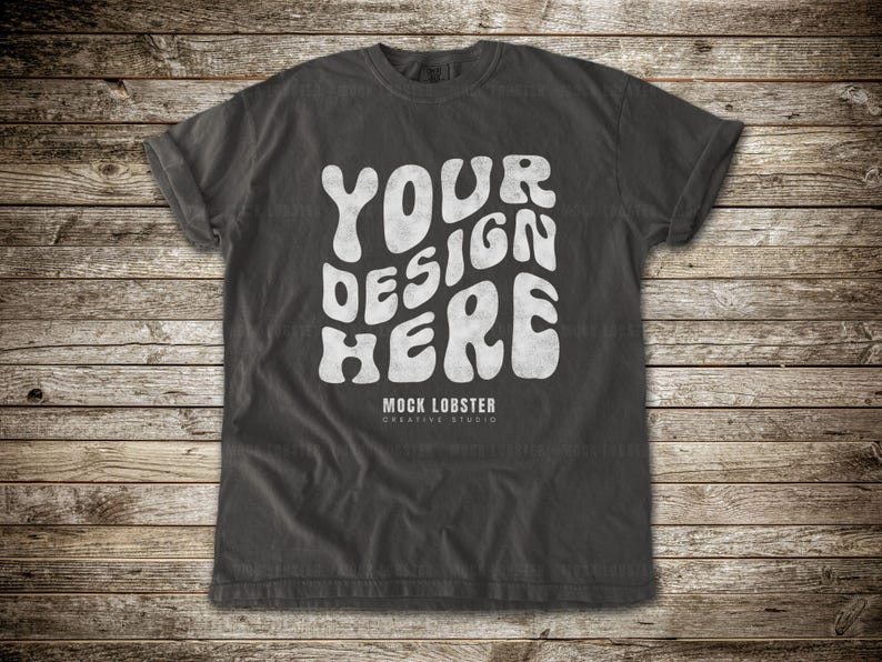 May include: Dark gray t-shirt with the text "YOUR DESIGN HERE" in a white, wavy font. The text "MOCK LOBSTER CREATIVE STUDIO" is below the main text. The shirt is displayed on a wooden background.