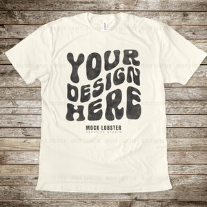 Op de afbeelding: Een wit T-shirt met een zwarte grafische afbeelding waarop "YOUR DESIGN HERE" staat in een retro, psychedelisch lettertype. De tekst is licht beschadigd, wat het een vintage uitstraling geeft. Het shirt ligt plat op een houten oppervlak.