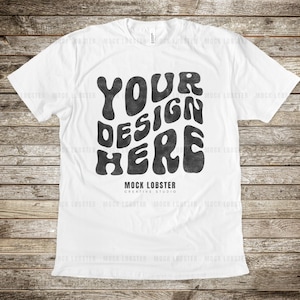 Könnte beinhalten: Weißes T-Shirt mit einem schwarzen Grafikdesign, auf dem in einer retro-psychedelischen Schrift "YOUR DESIGN HERE" steht. Der Text ist leicht verblasst und texturiert, was ihm einen Vintage-Look verleiht. Das Shirt liegt flach auf einer Holzoberfläche.