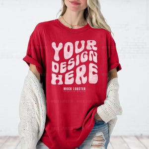 May include: A red t-shirt featuring a white graphic that reads "YOUR DESIGN HERE" in a retro typeface. The shirt is being modelled by a person wearing a white cardigan and blue jeans.