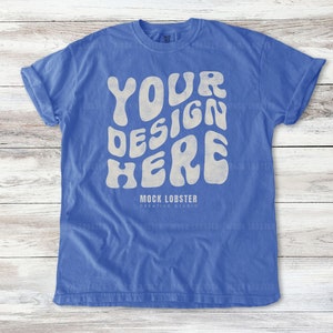May include: A blue t-shirt with a white graphic that says "YOUR DESIGN HERE" in a retro font. The shirt is on a wooden background.