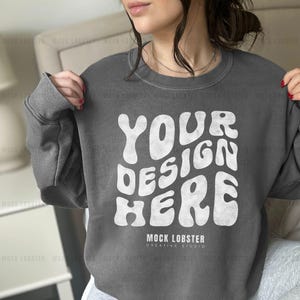 May include: A gray sweatshirt with the text "YOUR DESIGN HERE" in white, retro-style lettering. The text is centered on the sweatshirt and is surrounded by a white border. The sweatshirt has a crew neck and long sleeves.