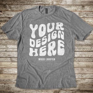 May include: A heather gray t-shirt with a white graphic that says "YOUR DESIGN HERE" in a retro font. The text is centered on the shirt and is surrounded by a white border. The shirt is laid flat on a wooden surface.