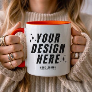 May include: A white ceramic mug with an orange rim and handle. The mug has the text "YOUR DESIGN HERE" in black lettering and the words "MOCK LOBSTER CREATIVE STUDIO" in smaller black lettering at the bottom.