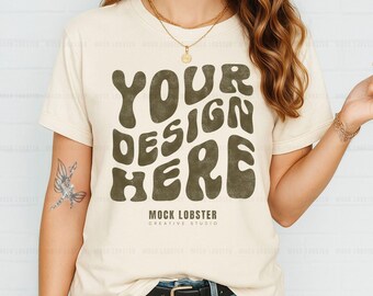 May include: A beige t-shirt with short sleeves and a crew neck. The shirt has a graphic design that says "Your Design Here" in a brown, retro font. The text "Mock Lobster Creative Studio" is printed below the graphic.