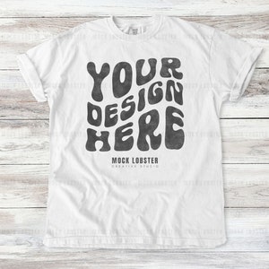 Könnte beinhalten: Weißes T-Shirt mit hochgekrempelten Ärmeln. Auf der Vorderseite des Shirts steht der Text "YOUR DESIGN HERE" in einer Retro-Schrift mit Wellen, darunter "MOCK LOBSTER CREATIVE STUDIO". Das Shirt wird vor einem hellen Holzgrund präsentiert.