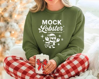May include: Olive green sweatshirt with the text "MOCK Lobster CREATIVE STUDIO" and a lobster graphic wearing a Santa hat and scarf. The person is wearing red and white plaid pajama pants and holding a mug with a red bow design.