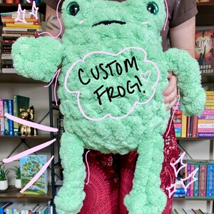 May include: A light green plush frog toy with a textured, fuzzy surface, large dark eyes, and a flat mouth. The frog features a pink hand-drawn cloud graphic on its chest with the text "CUSTOM FROG!" and a small heart symbol next to it.