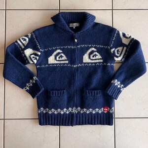May include: A navy blue zip-up knit jumper featuring a shawl collar and a white wave pattern across the chest and sleeves. The garment includes two front pockets, ribbed cuffs and hem, and a small red logo tag on the lower left side.