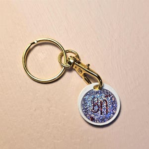 Könnte beinhalten: Ein goldfarbener Schlüsselanhänger mit einem runden, weiß umrandeten Anhänger. Der Anhänger hat einen blauen Glitzerhintergrund mit den Buchstaben "Bn" in Rot. Der Schlüsselanhänger enthält einen Karabiner und einen Schlüsselring.