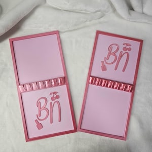 May include: Two pink rectangular signs with a raised border and a decorative band. Each sign has the letters "Bn" with a cherry and a bottle design. The signs are on a white surface.