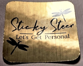 Może przedstawiać: Kwadratowa, złota naklejka z zaokrąglonymi rogami. Naklejka zawiera napis "Sticky Steer" kursywą, z napisem "Let's Get Personal" poniżej. Obecne są również dwie sylwetki ważek.