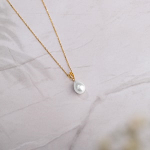 May include: A gold-colored chain necklace with a pear-shaped, white pearl pendant. The necklace is displayed on a white and gray marble surface. The pendant is a teardrop shape.