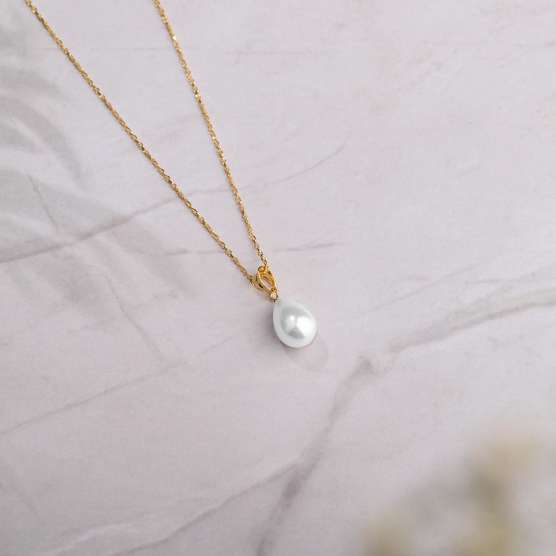 May include: A gold-colored chain necklace with a pear-shaped, white pearl pendant. The necklace is displayed on a white and gray marble surface. The pendant is a teardrop shape.