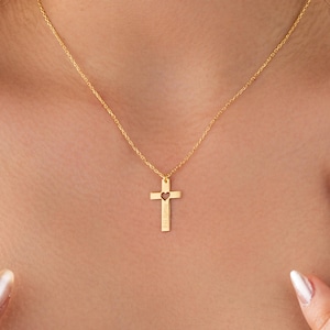 May include: A gold-colored necklace featuring a cross pendant with a heart-shaped cutout. The cross hangs from a delicate chain. The pendant has a small inscription at the bottom. The necklace is displayed against a skin-toned background.