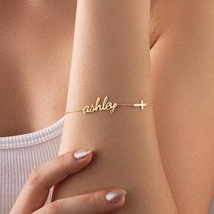 May include: A delicate gold bracelet featuring the name "ashley" in cursive script. A small cross charm is attached to the bracelet. The bracelet is worn on a person's wrist.