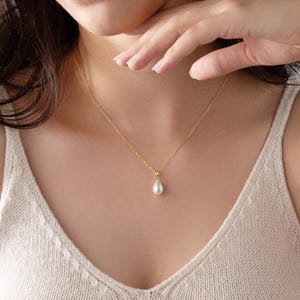 May include: A gold-colored necklace with a teardrop-shaped pearl pendant. The necklace is delicate and hangs on a person's neck, against a neutral-colored top. The pearl is white and has a smooth surface.
