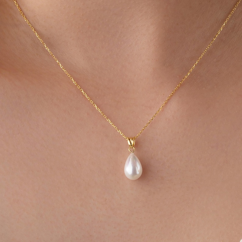 May include: A delicate gold necklace featuring a pear-shaped, white pearl pendant. The chain is a fine, gold link, and the pendant hangs gracefully. The necklace is shown against a skin-toned background, highlighting the jewelry's elegance.