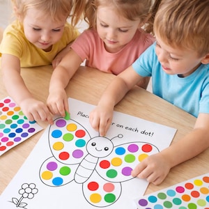 Könnte beinhalten: Drei kleine Kinder platzieren bunte Aufkleber auf einem Schmetterling-Aktivitätsblatt. Das Blatt hat eine Zeichnung eines Schmetterlings mit Kreisen für die Aufkleber. Der Text "Place...er on each dot!" ist sichtbar. Bunte Aufkleberbögen liegen ebenfalls auf dem Tisch.