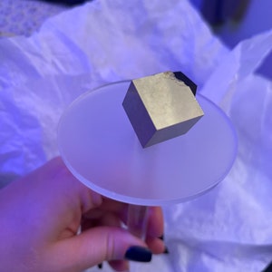 May include: A silver cube-shaped crystal sits on a clear glass pedestal. The crystal is resting on a white surface.