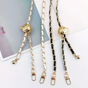 Pode incluir: Três alças de bolsa de corrente dourada com fecho dourado e um detalhe de bola dourada. As correntes são brancas, pretas e brancas e pretas.
