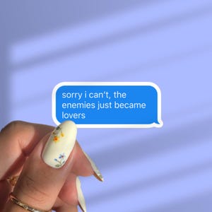 May include: A blue and white sticker with the text "sorry i can't, the enemies just became lovers" on a light purple background. The sticker is in the shape of a text message bubble.