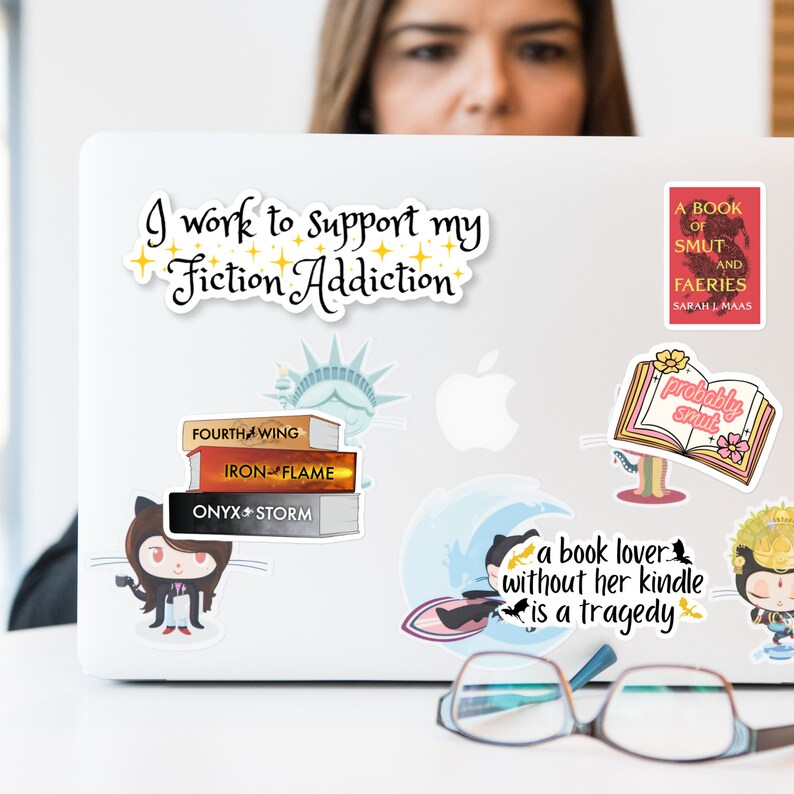 Op de afbeelding: Een laptop met verschillende stickers erop. De stickers bevatten een stapel boeken met de titels "Fourth Wing", "Iron Flame" en "Onyx Storm", een cartoon van een kat met een kopje koffie, een boek met de tekst "probably smut", een boek met de tekst "A Book of Smut and Faeries", een cartoon van een kat die op een golf surft met de tekst "a book lover without her kindle is a tragedy", en een cartoon van een kat met een kroon. De laptop heeft ook een sticker met de tekst "I work to support my Fiction Addiction".