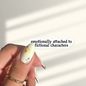 Puede incluir: Pegatina blanca con el texto "emocionalmente apegado a personajes ficticios" sobre un fondo blanco. Una mano con uñas de arte floral sostiene la pegatina.