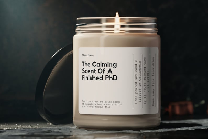 K&ouml;nnte beinhalten: Eine durchsichtige Glaskerze in einem Glas mit einem wei&szlig;en Etikett, auf dem "The Calming Scent Of A Finished PhD" steht. Die Kerze ist aus handgegossenem Sojawachs und in Kalifornien hergestellt. Sie hat eine Brenndauer von 50 bis 60 Stunden und wiegt 255 g (9 oz).