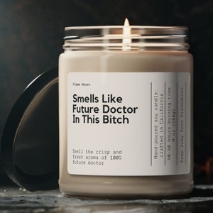 Puede incluir: Una vela de soja encendida en un tarro de cristal con tapa plateada. La etiqueta dice "Smells Like Future Doctor In This Bitch". La vela es de 255g y está vertida a mano en California.