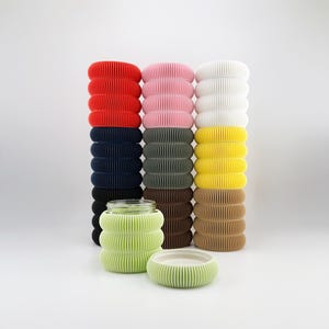 Puede incluir: Una colección de objetos cilíndricos acanalados de colores, incluyendo rojo, rosa, blanco, azul marino, verde oliva, amarillo y marrón. Un tarro verde y un cuenco pequeño a juego están en primer plano. Los objetos están apilados en columnas.