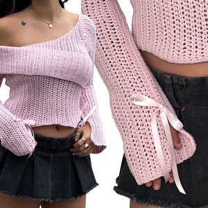 Könnte beinhalten: Ein rosa gehäkelter Pullover mit einem lockeren, off-shoulder-Ausschnitt und langen Ärmeln. Die Ärmel haben ein Bindedetail am Handgelenk. Der Pullover ist kurz und zeigt ein Stück des Bauches des Models.