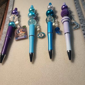 Op de afbeelding: Vier decoratieve pennen met kralen tops en bedels. De pennen hebben een verscheidenheid aan kleuren, waaronder paars, lichtblauw en wit. Elke pen heeft een zilverkleurige metalen punt en een unieke bedel, zoals een foto of een boze oog.