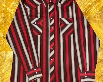 May include: A long-sleeved, red and grey striped Western shirt with a pointed collar, button-down front, and two chest pockets. The shirt has the Wrangler brand label on the left chest.