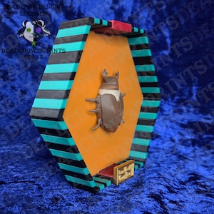 Puede incluir: Una vitrina geom&eacute;trica con una figura de escarabajo marr&oacute;n sobre un fondo naranja. La vitrina tiene detalles en negro, turquesa y rojo. El texto "DEADCROWDESIGNS SOCIALS" y "DEADCROW3DPRINTS STORE" son visibles.