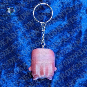 May include: A keychain featuring a metallic silver split ring and a short chain attached to a small, pink, rectangular-shaped charm with a textured, slightly shimmering surface and a rounded top edge, suspended vertically against a blue background.