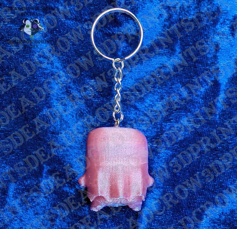 May include: A keychain featuring a metallic silver split ring and a short chain attached to a small, pink, rectangular-shaped charm with a textured, slightly shimmering surface and a rounded top edge, suspended vertically against a blue background.