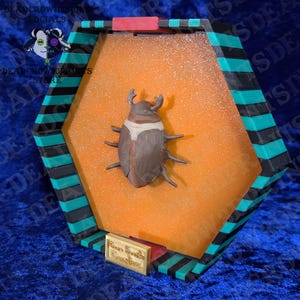 Puede incluir: Una vitrina decorativa hexagonal con una escultura de escarabajo marr&oacute;n y blanco. La caja tiene un borde negro, verde azulado y rojo. Una peque&ntilde;a placa dorada en la parte inferior dice "Your Beetle Creations".