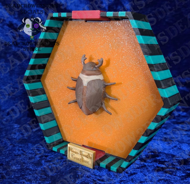 Puede incluir: Una vitrina decorativa hexagonal con una escultura de escarabajo marr&oacute;n y blanco. La caja tiene un borde negro, verde azulado y rojo. Una peque&ntilde;a placa dorada en la parte inferior dice "Your Beetle Creations".