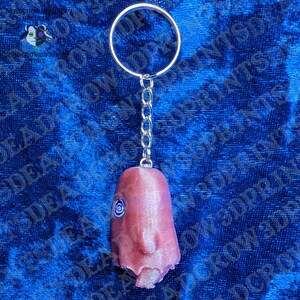 May include: A keychain featuring a metallic split ring and a short chain attached to a pink, textured, three-dimensional figure. The figure has a smooth, rounded top, a prominent nose, and a single spiral eye detail on the side of its head.