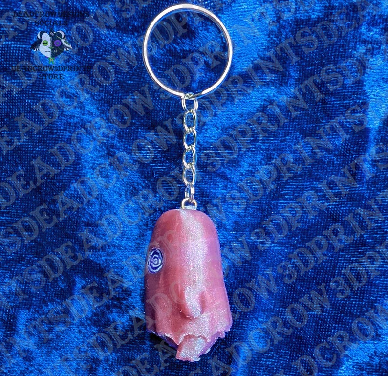 May include: A keychain featuring a metallic split ring and a short chain attached to a pink, textured, three-dimensional figure. The figure has a smooth, rounded top, a prominent nose, and a single spiral eye detail on the side of its head.